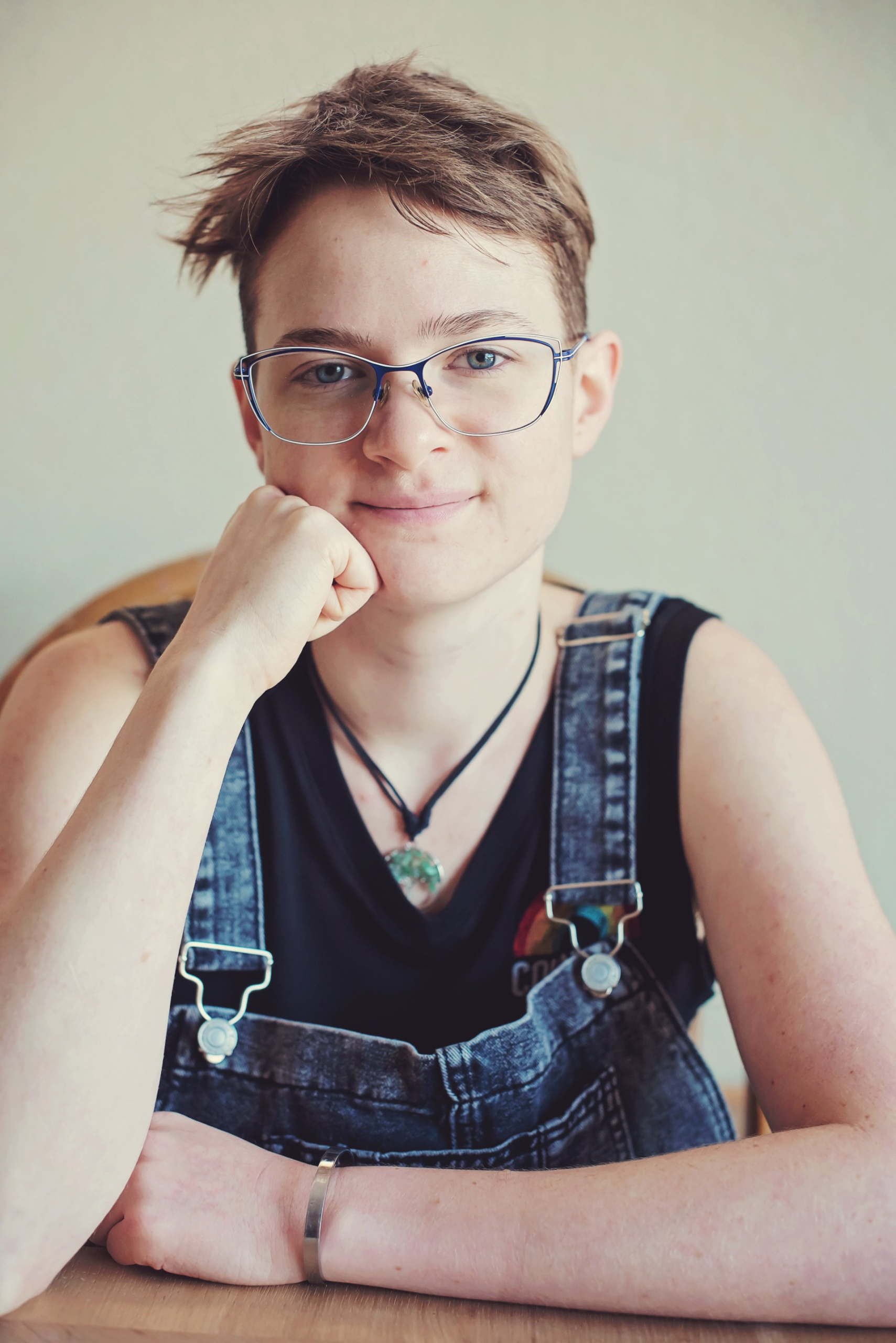 Kayda sitting at a table. They are white with short brown hair. They are wearing overalls, a black tank top, glasses, a metal bracelet, and a tree necklace.