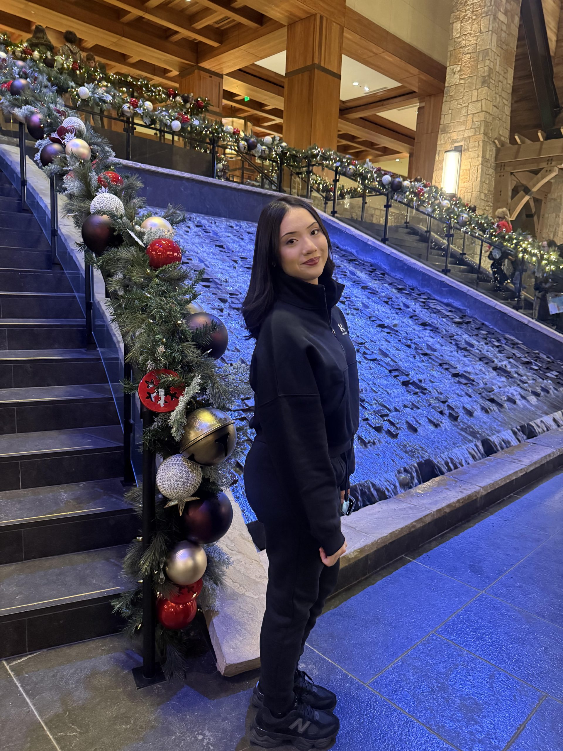Alyssa standing in front of two beautiful staircases covered in Christmas decorations with water flowing in the middle. She has long black hair and is wearing all black.