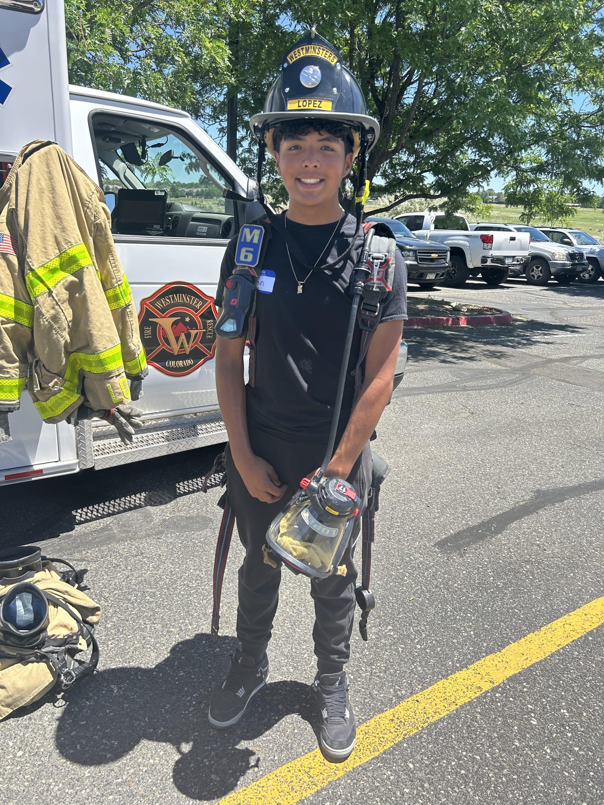 Gian standing in front of a Westminster Fire Rescue truck smiling and wearing a firefighter helmet and holding a respirator. He has short black hair and brown skin.
