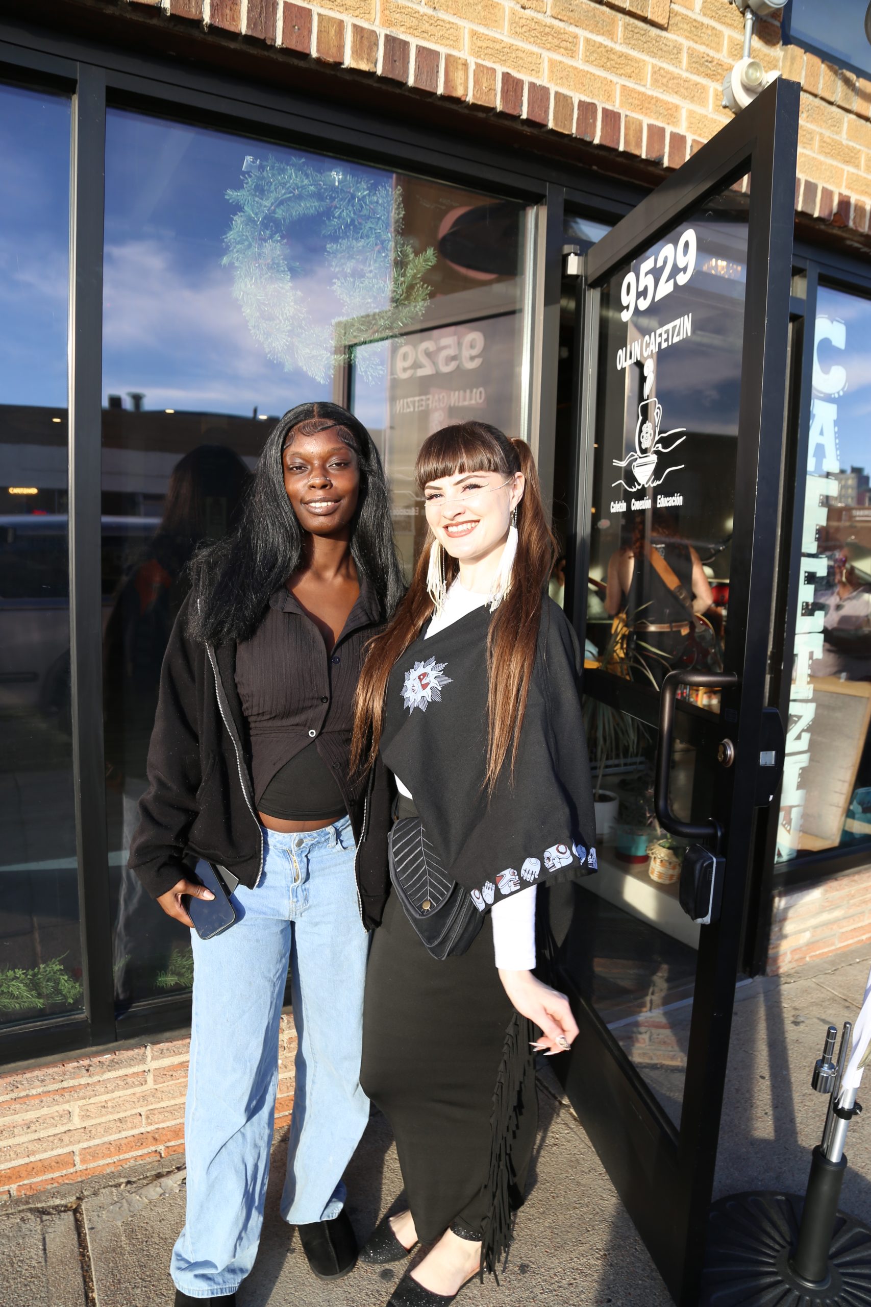 Janae is a black woman with long black hair. She is wearing a black top and a black jacket with the zipper unzipped. She is also wearing bright blue jeans and black shoes. She is standing next to Sedona who has light skin and long brown hair. She is wearing white jewelry with small pearls that goes across the bridge of her nose and behind her ears. She is also wearing a black top, black skirt, and black shoes. They are both standing in front of Ollin Cafetzin in Denver on a sunny day.