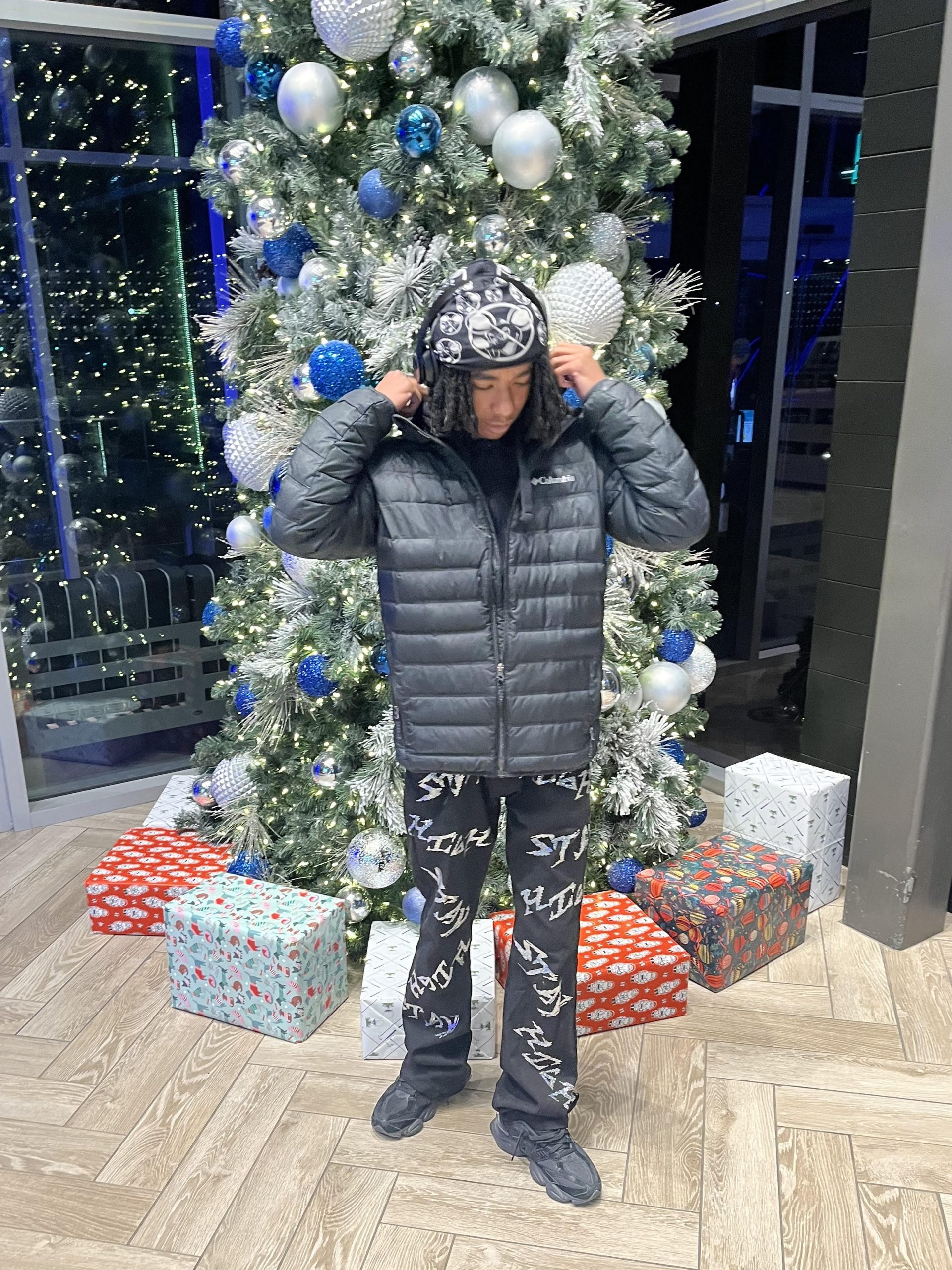 Lijah standing in front of a Christmas tree. He is black with chin length dreads, and he’s wearing headphones, a black coat, pants, and shoes.