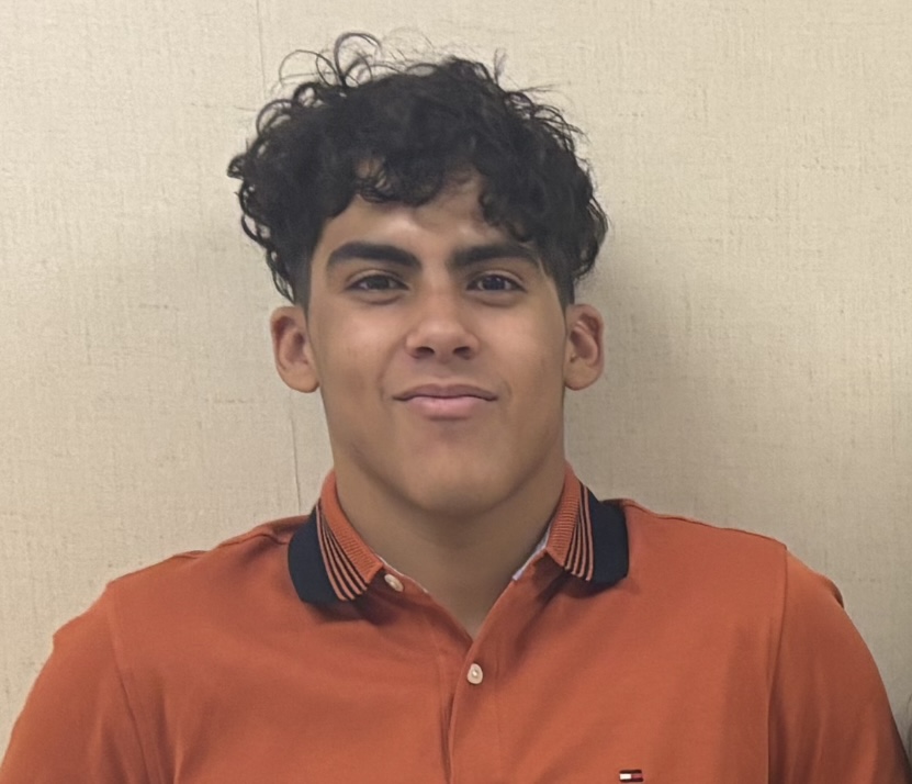 Headshot photo of Muty. He is standing against a tan wall wearing a bright orange shirt. He has brown skin and hair. His hair has loose curls that are a few inches long. He is quite muscular and has broad shoulders, but he looks very kind and caring.