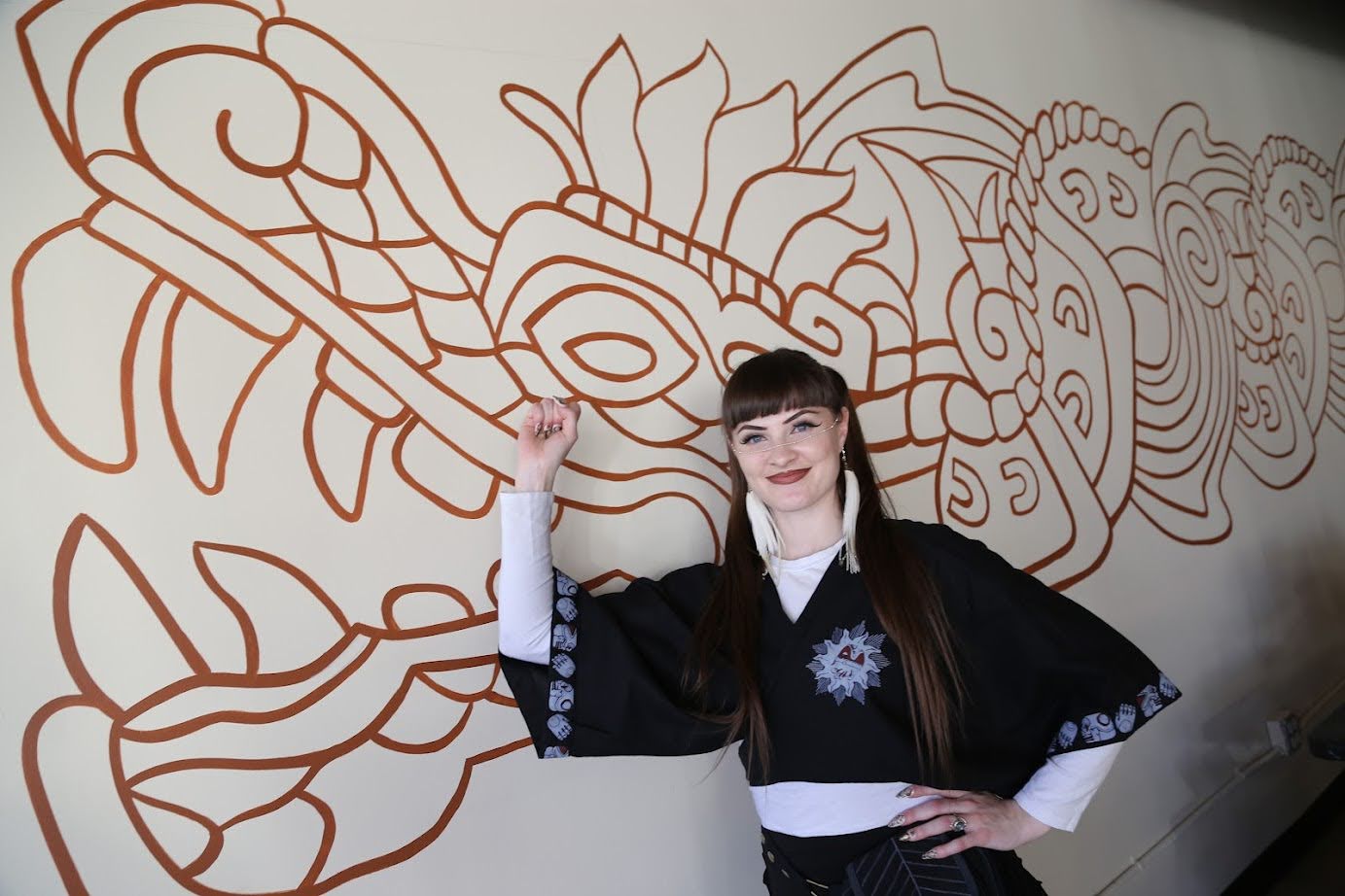 Sedona standing in front of a Chinese dragon mural. Sedona has long brown hair in pig tails, light skin, and is wearing a white long sleeve shirt with a black short sleeve shirt on top, black pants, and jewelry that goes across her nose and behind her ears with a white tassel at either end.