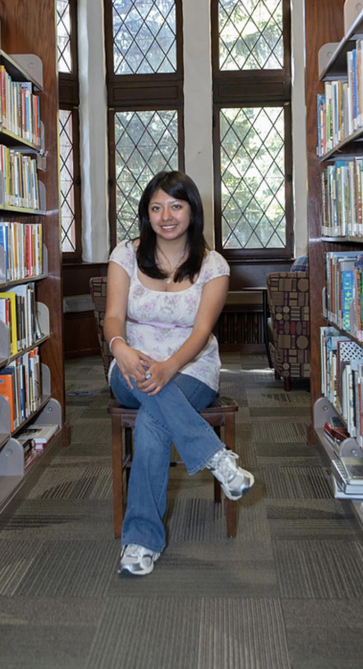 Silvia sitting in a library while smiling. She has long dark hair, and is wearing a necklace, white top with a floral pattern, and jeans.