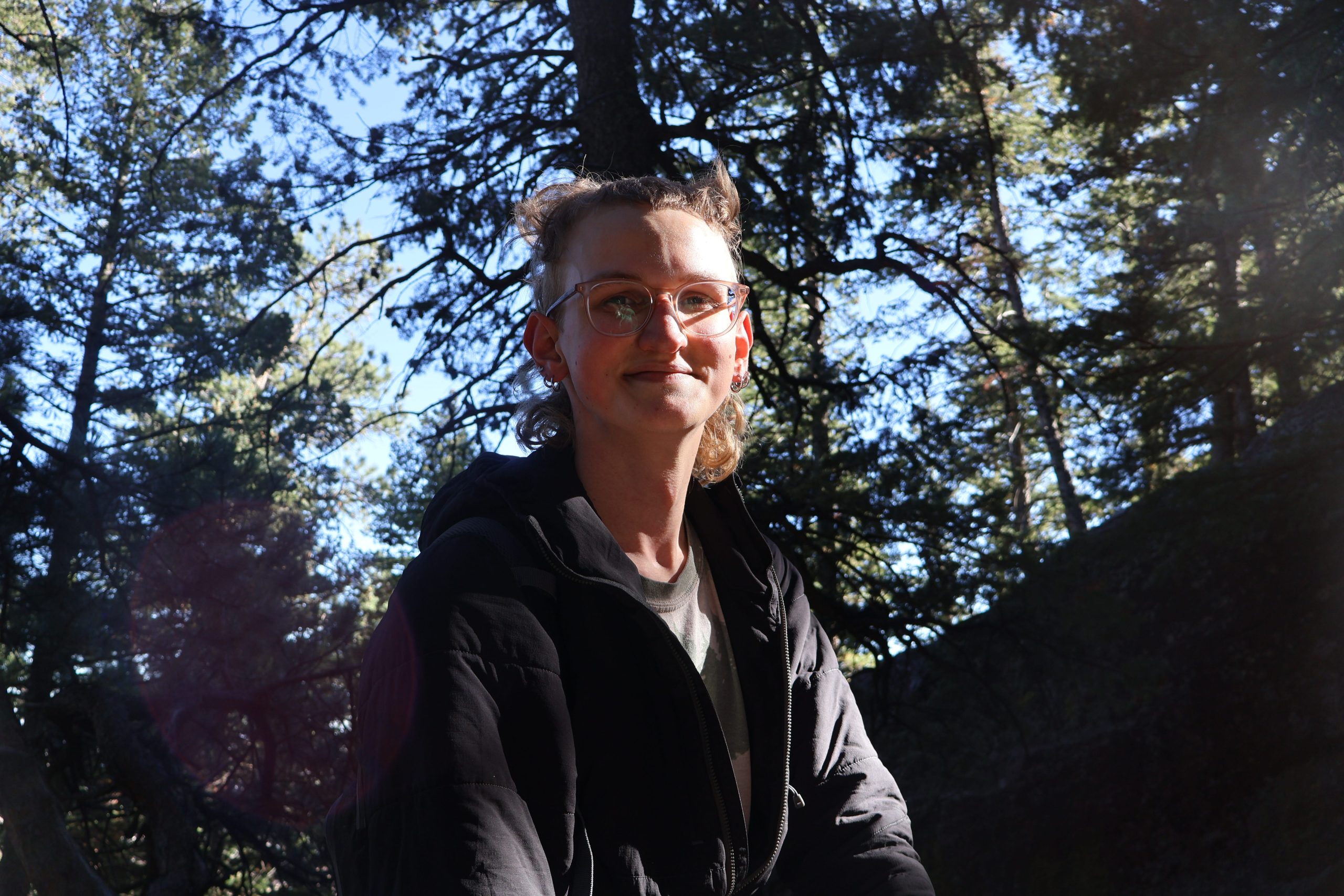 Sybil in the woods. They have light skin, curly blond hair that is nearly shoulder length, and they are wearing glasses, a gray shirt, and black jacket.