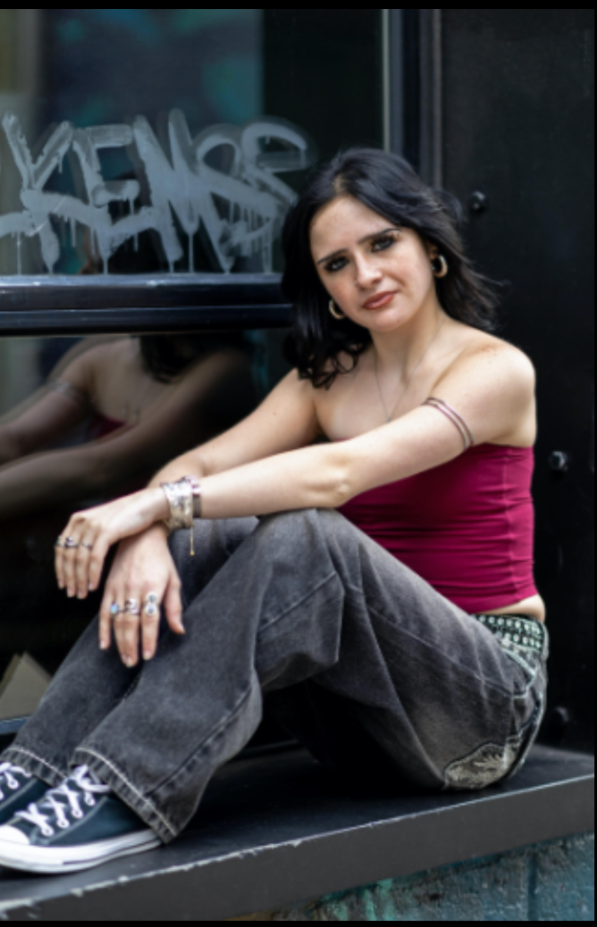 Tay sitting on a window ledge outside. They have light skin and black, shoulder length hair. She is wearing jeans, a magenta, sleeveless top, and various jewelry.